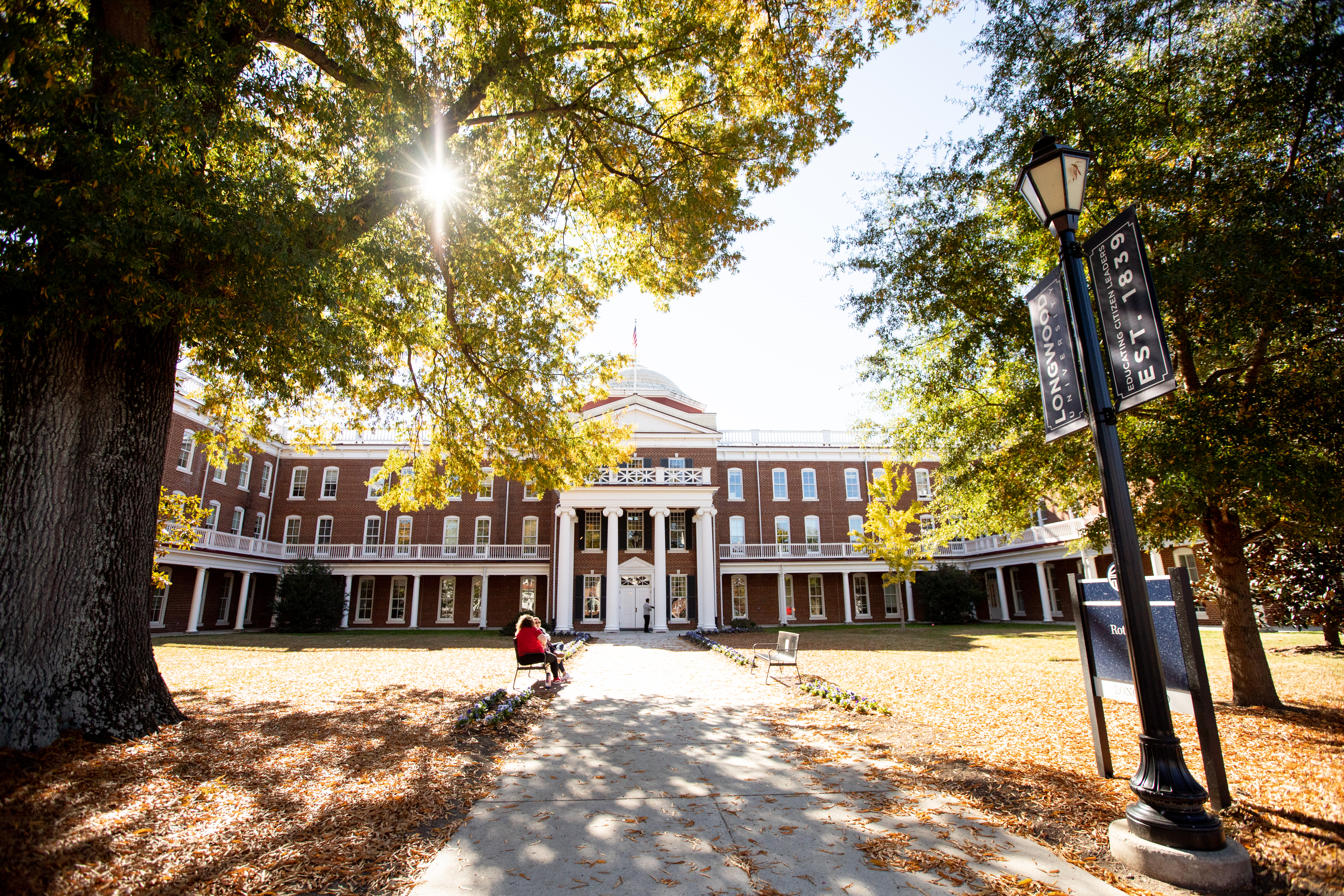 Rotunda with sun peeking through the trees and two students sitting on a bench out front - Sunlight filters through autumn trees in front of Rotunda Hall. The red-brick building with white columns stands at the end of a leaf-covered walkway. A campus banner reading “Longwood EST. 1839” hangs from a lamppost. Students sit on benches in the warm fall afternoon light.