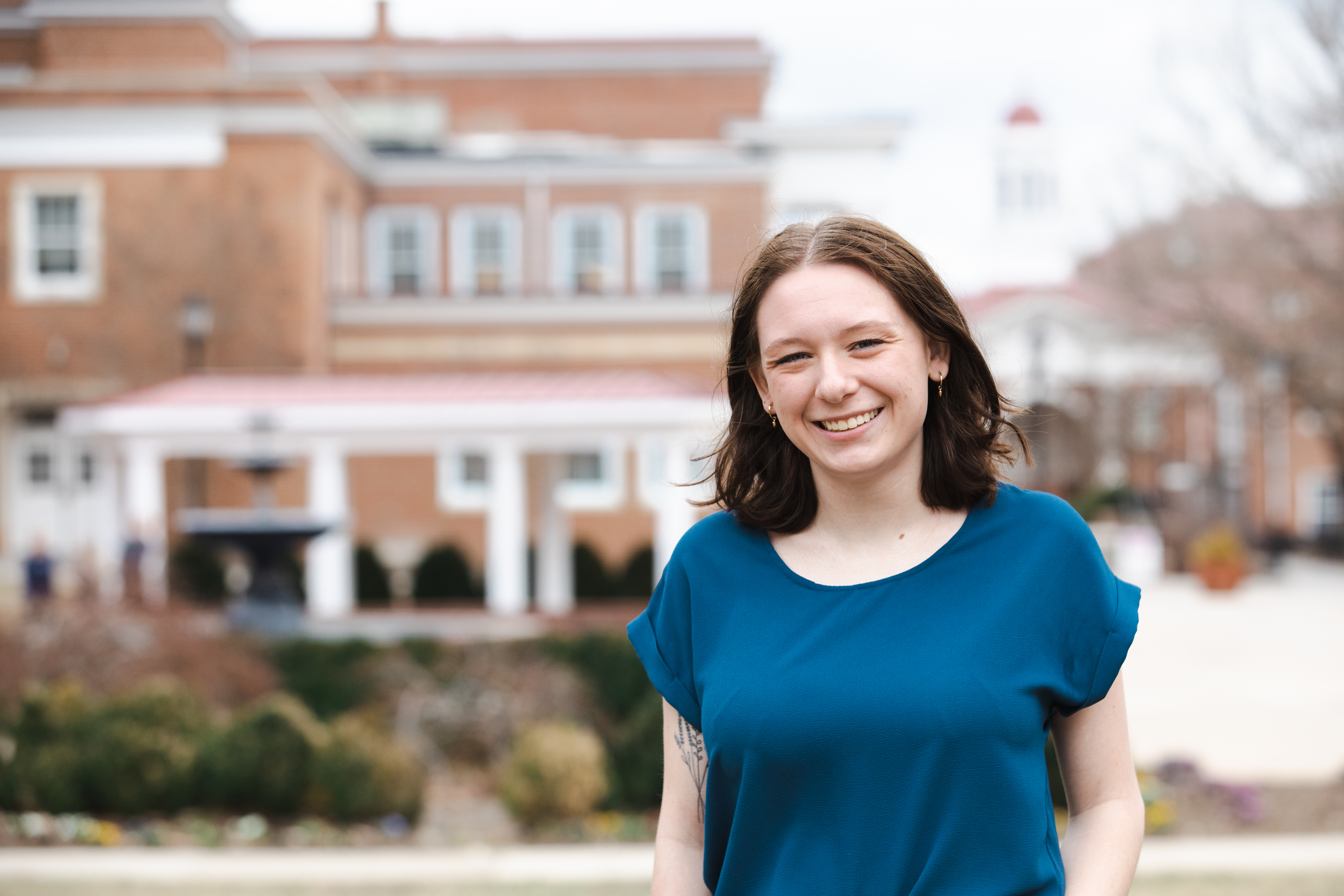 Emily Robertson ’24 - A student smiles while standing outdoors on campus. A brick academic building with white columns is visible behind her. Landscaping and a fountain appear in the background. The image is a relaxed campus portrait.