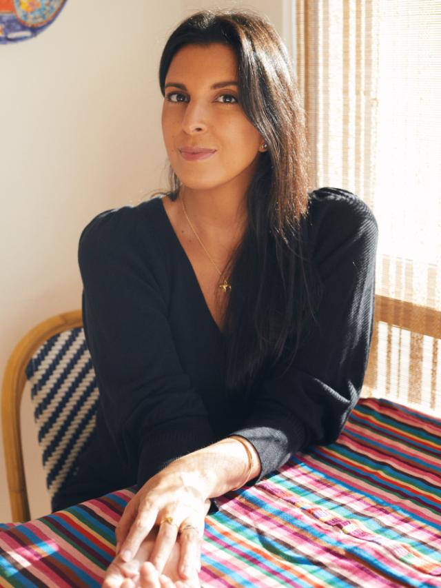 Patricia Engel, credit: Elliott and Erick Jimenez - Portrait of a woman seated at a table in soft sunlight. She wears a black top and a delicate gold necklace. Her hands rest on a colorful striped tablecloth. Light filters through sheer curtains behind her.