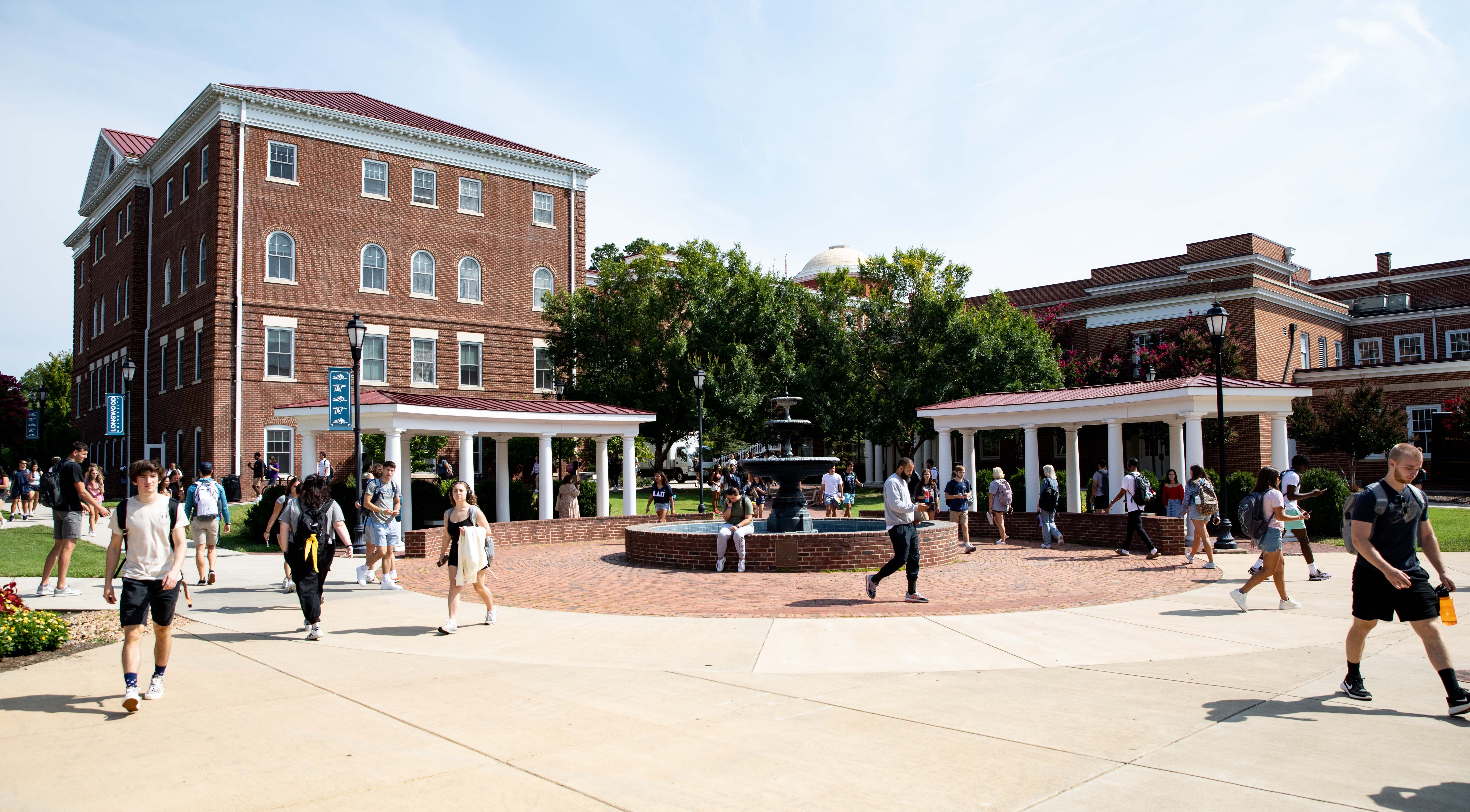 Students walking across Beale Plaza - Students walk across campus near a brick fountain and white-columned pergolas. Red-brick academic buildings frame the courtyard under a clear sky. Some students carry backpacks and water bottles as they head to class. Green lawns and trees surround the busy quad.