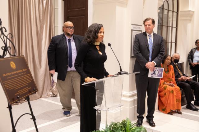 Lori Isaac speaks at the Dedication of Allen Hall - A woman speaks at a podium during a dedication ceremony. A plaque honoring Dr. Edna Allen Bledsoe is displayed on an easel nearby. Officials stand behind her while guests are seated along the wall. The event takes place in a bright hall with columns and arched windows.