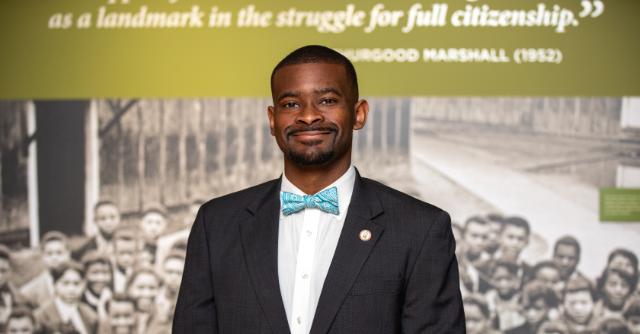 Cainan Townsend ’15, M.S. ’20 - A man in a dark suit and teal bow tie stands smiling for a portrait. He wears a lapel pin and stands in front of a historical exhibit display. A blurred black-and-white photograph and quote appear in the background. The image presents a professional headshot in a museum setting.
