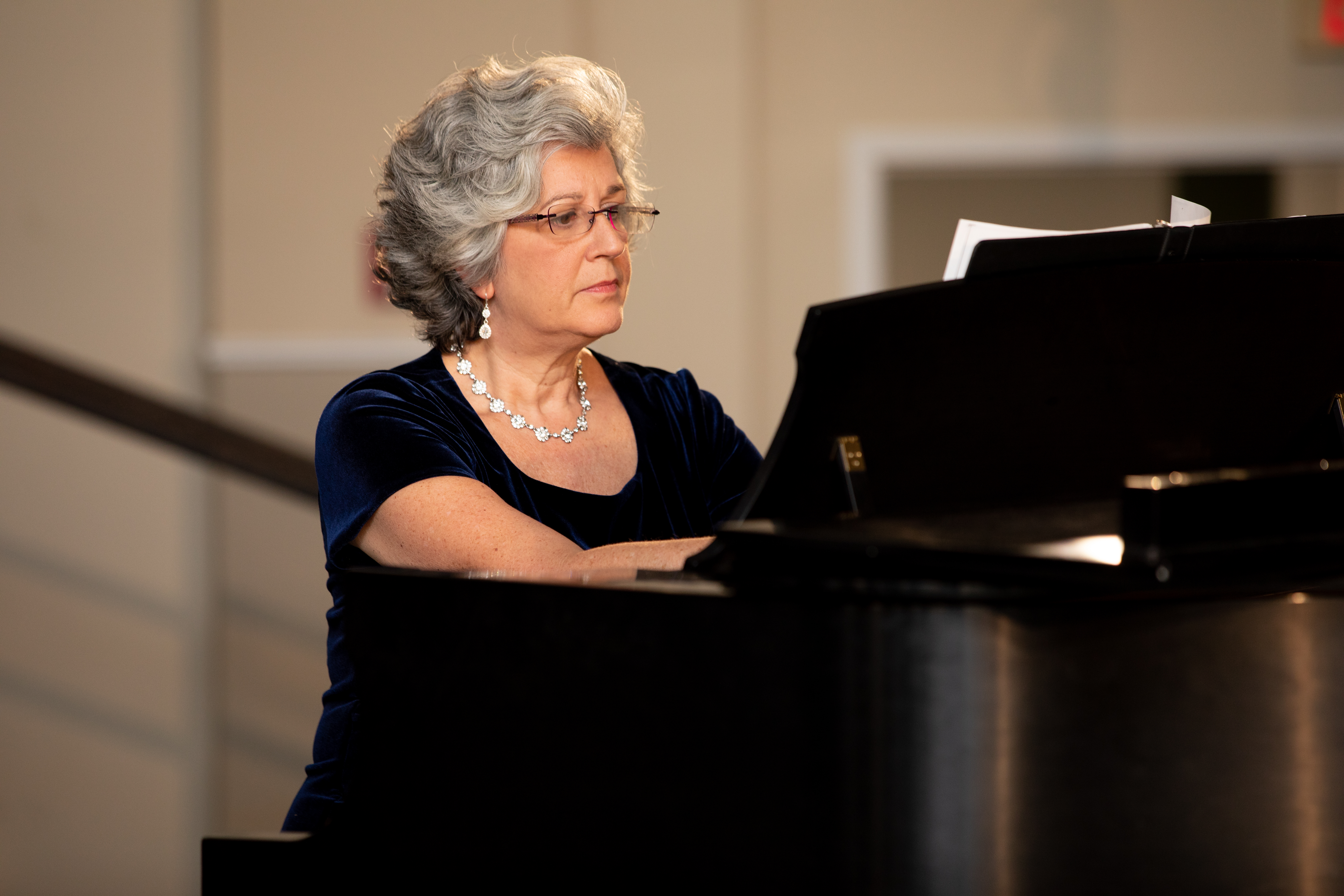 Dr, Lisa Kinzer during a concert performance - A pianist performs at a grand piano during a holiday concert. She wears a dark blue dress with a necklace and earrings. Sheet music rests open on the piano stand. Soft lighting and a recital hall setting frame the performance.