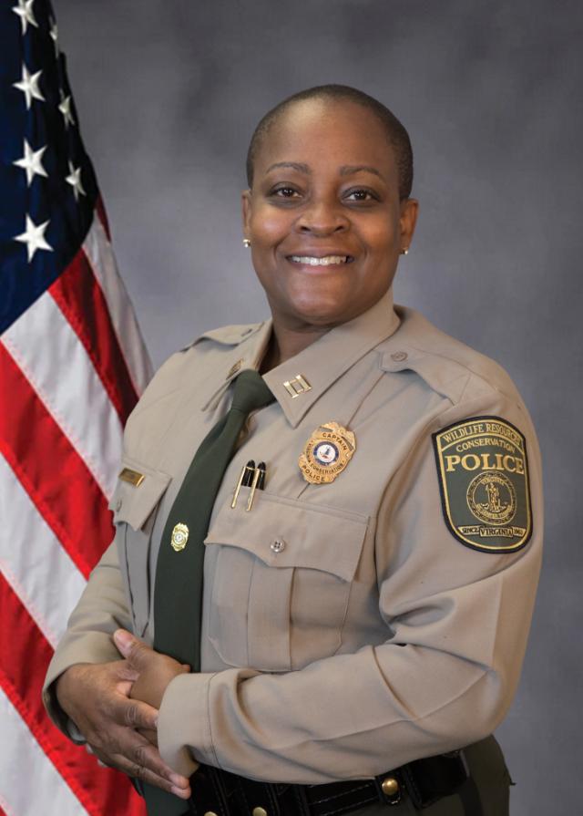Angela Comer, new chief of the Longwood University Police Department - A uniformed Wildlife Resources Conservation Police officer poses for a formal portrait. She wears a tan dress uniform with badge, insignia, and a green tie. An American flag stands behind her against a neutral studio backdrop. She smiles with her hands folded in front of her.