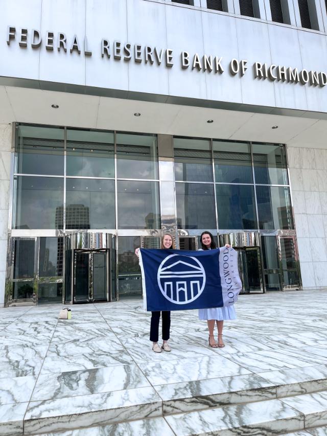 Katya Mayer ’24 and Charlotte Austin ’23 - Two women stand on a marble plaza holding a Longwood University flag. They are positioned in front of the Federal Reserve Bank of Richmond building. The building’s name is displayed above tall glass windows and revolving doors. City buildings reflect in the glass facade behind them.