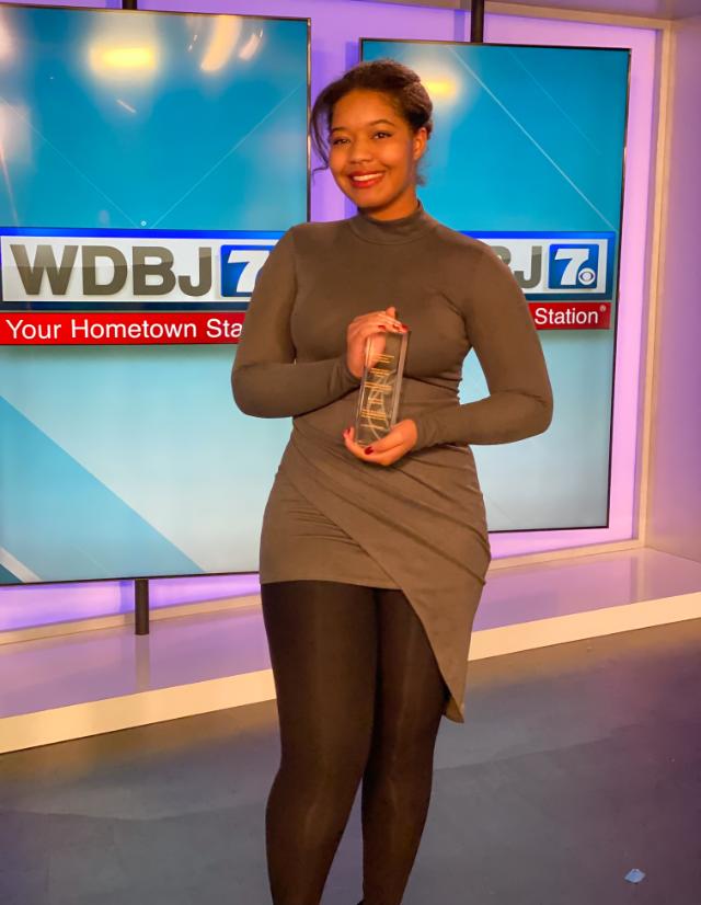 Makayla Jennings ‘22 - A woman stands in a television studio holding a clear award plaque. She smiles at the camera in front of screens displaying “WDBJ7” and “Your Hometown Station.” She wears a long-sleeve dress with black tights and heels. The studio lighting casts a soft purple and blue glow in the background.
