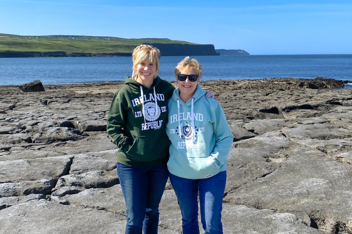 Dr. Kim Little and Lisa Minor in Ireland during the study abroad experience - Two women stand together on a rocky shoreline with the ocean behind them. They wear hoodies that read “Ireland Republic” and smile at the camera. Green cliffs and calm blue water stretch across the background under a clear sky. The photo captures a bright coastal scene during a visit to Ireland.
