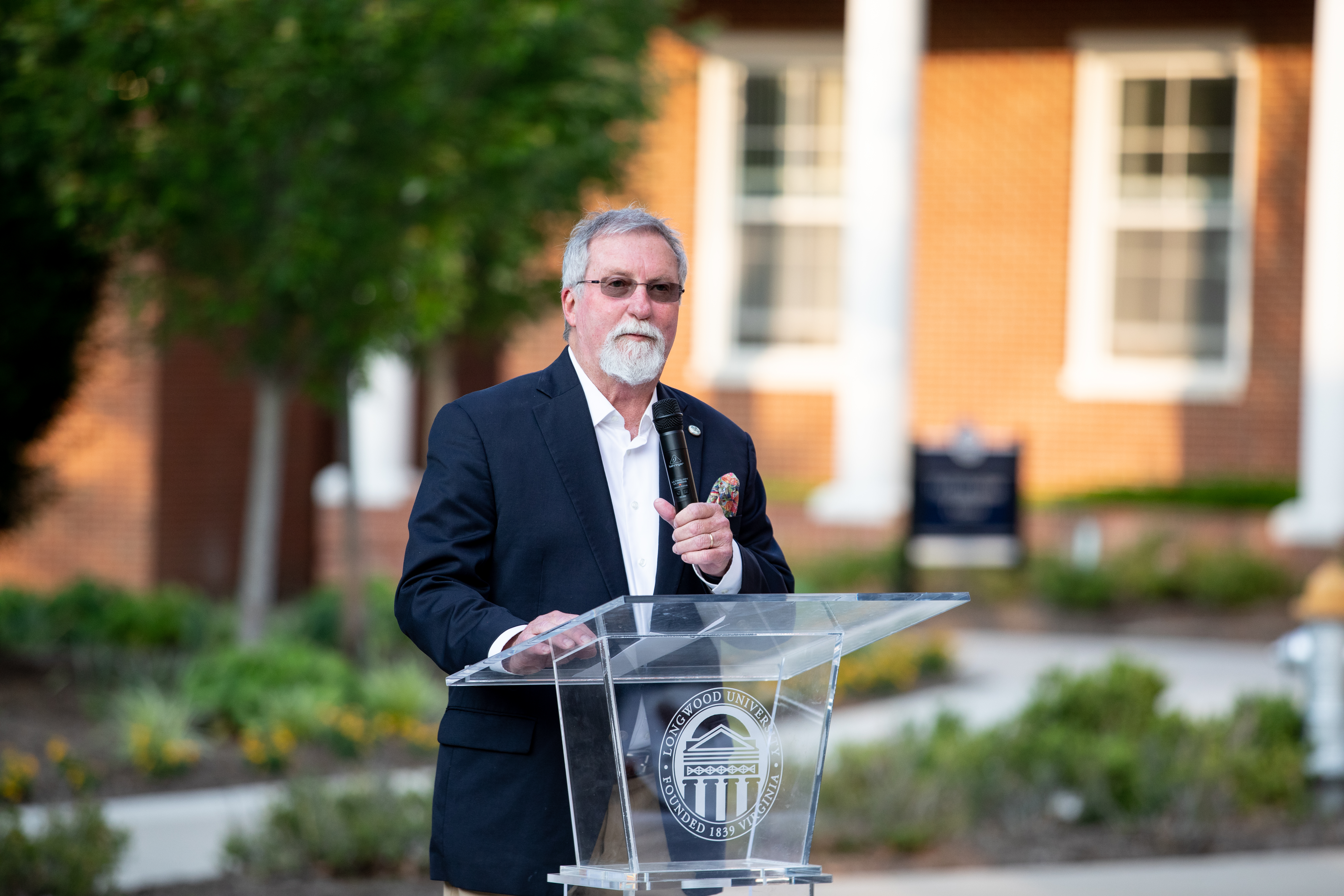 Dr. Ken Perkins speaking at a retirement celebration held in his honor last month - A man in a dark suit speaks into a microphone at a clear podium outdoors. The podium displays the Longwood University seal. Red-brick buildings and white columns stand in the background. He appears to be addressing a crowd during a campus event.