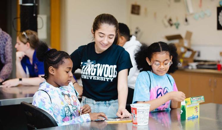 Longwood student working with school children during a Lancer for the Day event - A Longwood University student in a green T-shirt works with two young girls at a classroom table. They focus on a hands-on activity with paper and small materials spread out in front of them. One child looks at the project while the other reaches into a small box. The image shows mentoring and learning during a campu