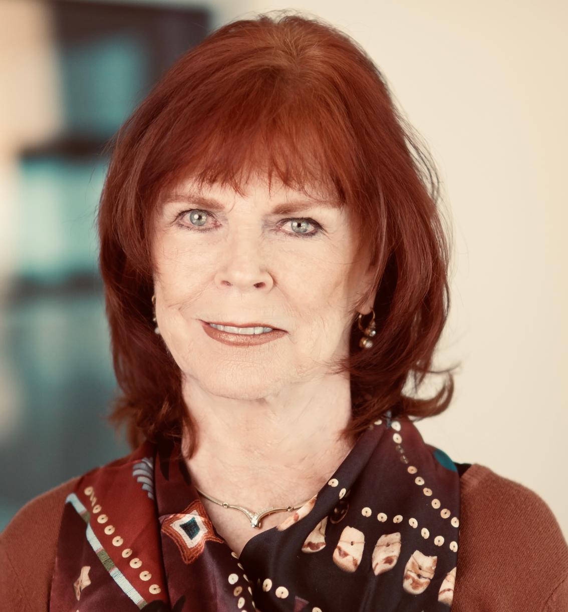 Lynn Gardner Heffron ’67 - Headshot of a woman with shoulder-length red hair and blue eyes. She wears a patterned scarf and small earrings. Her expression is calm as she faces the camera. The background is softly blurred indoors.