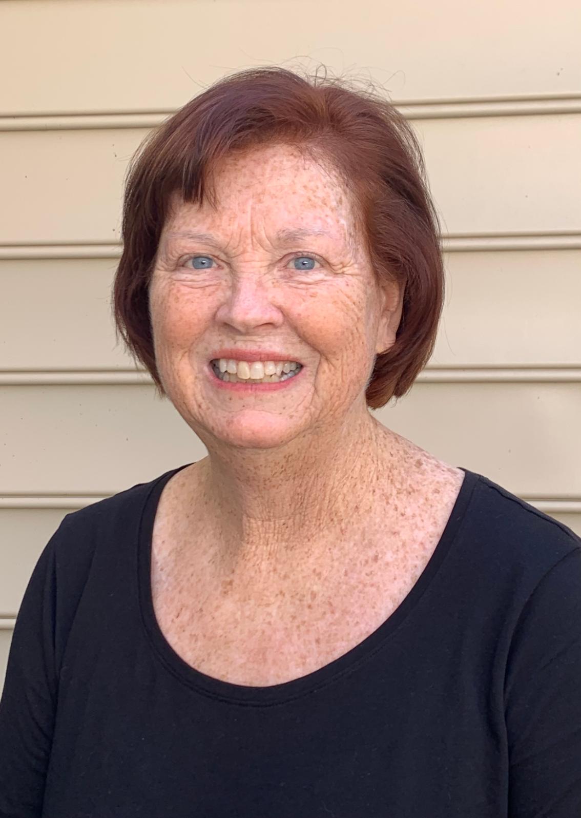 Deana McGuire Buck ’83 - Headshot of a smiling woman with short auburn hair and blue eyes. She wears a simple black top. The portrait is taken outdoors against a light tan siding background. She faces the camera with a warm expression.