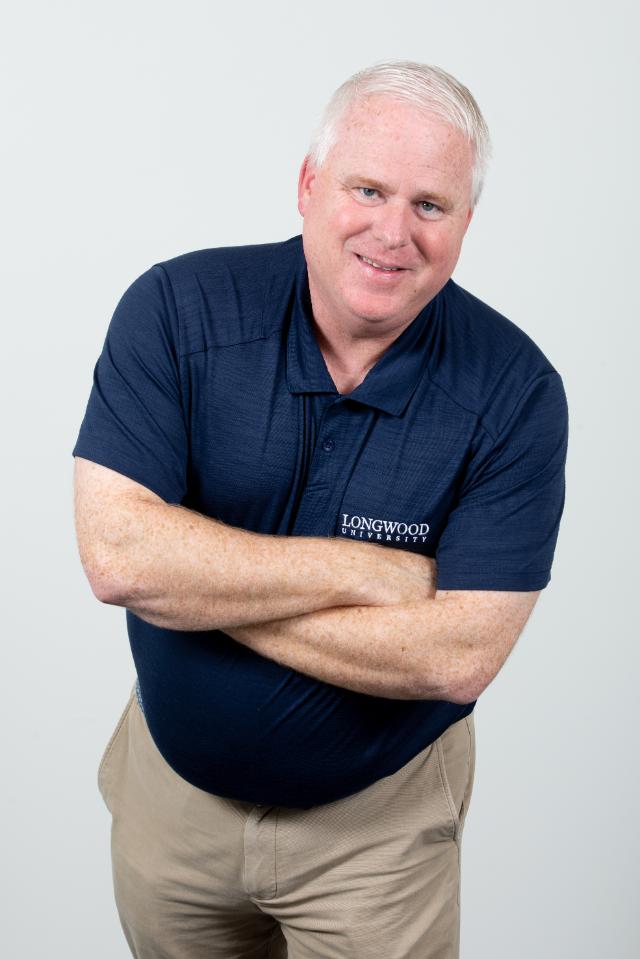Jason “Ferg” Ferguson - Portrait of a man with short white hair smiling at the camera. He stands with arms crossed against a light gray background. He wears a navy polo embroidered with “Longwood University.” Khaki pants complete the professional, casual look.