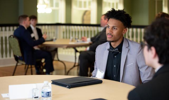 Business students engage in mock interviews as part of a course to develop workplace skills - A student in a light gray suit and bow tie sits at a table during a mock interview. He listens attentively to an interviewer across from him with a portfolio on the table. Other pairs of participants are visible in conversation in the background. The image captures a professional development event in a ca