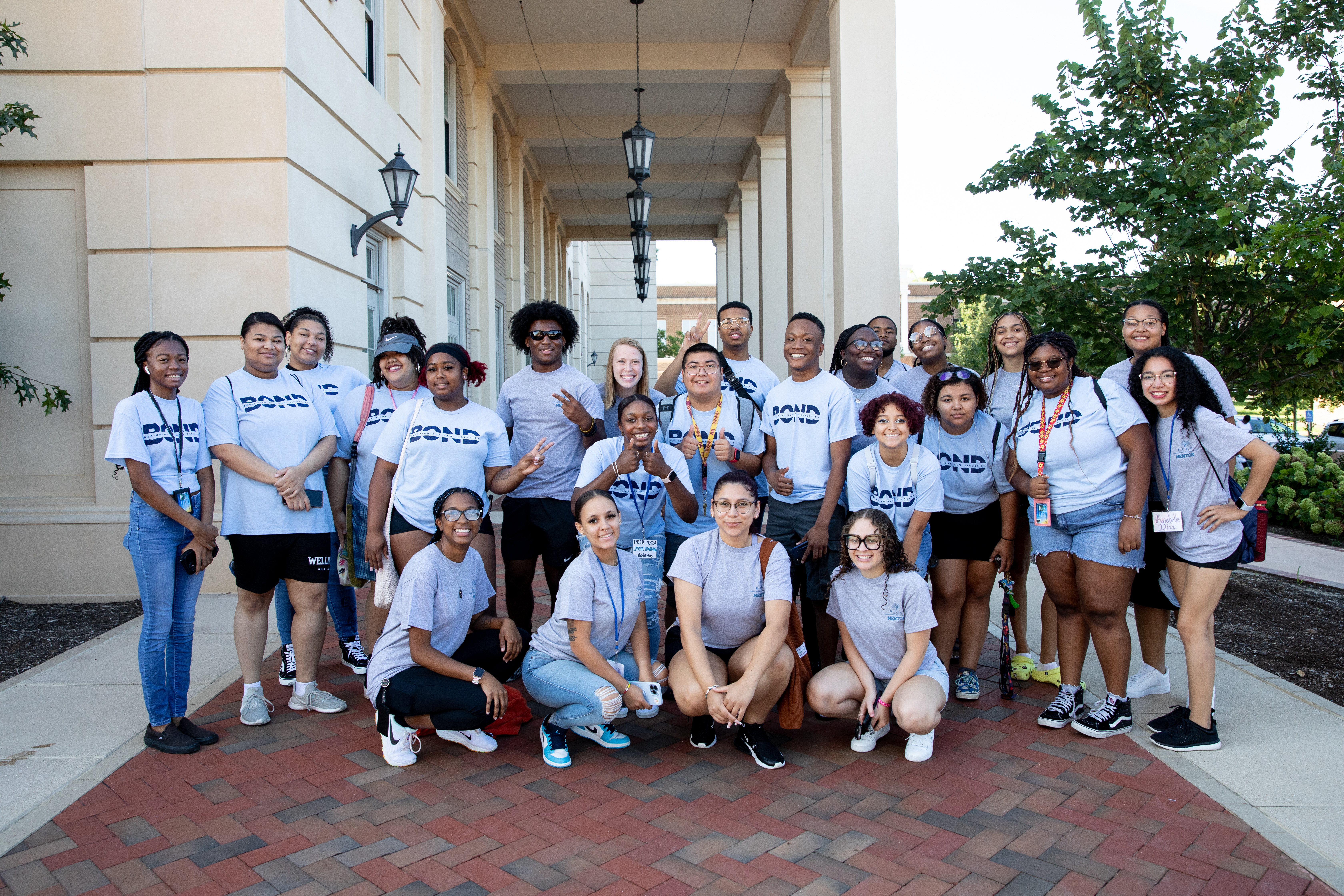 BOND students and mentors posing outside Johns and Moss - A large group of students wearing matching gray T-shirts pose together outdoors. They stand in front of a campus building with tall columns and hanging lanterns. Some students kneel in the front row while others stand behind them, smiling. Trees and brick walkways frame the bright, sunny campus setting.