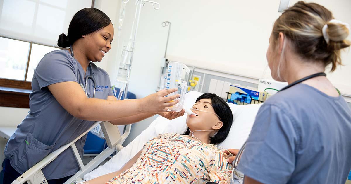 Nursing Students tending to a patient in the simulation lab - Two nursing students practice clinical skills in a simulation lab. One adjusts an IV fluid bag while the other checks equipment. A medical training mannequin lies in a hospital bed. Monitoring devices and clinical tools surround the bedside.