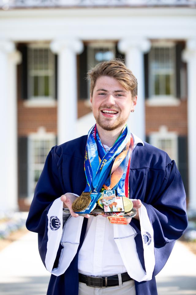 Jacob Barker ’23 - A graduate in a navy gown stands in front of a red brick campus building with white columns. He smiles while holding a collection of medals in his hands. Colorful ribbons hang around his neck from multiple races and competitions. The portrait highlights his academic achievement and athletic accomplishments.
