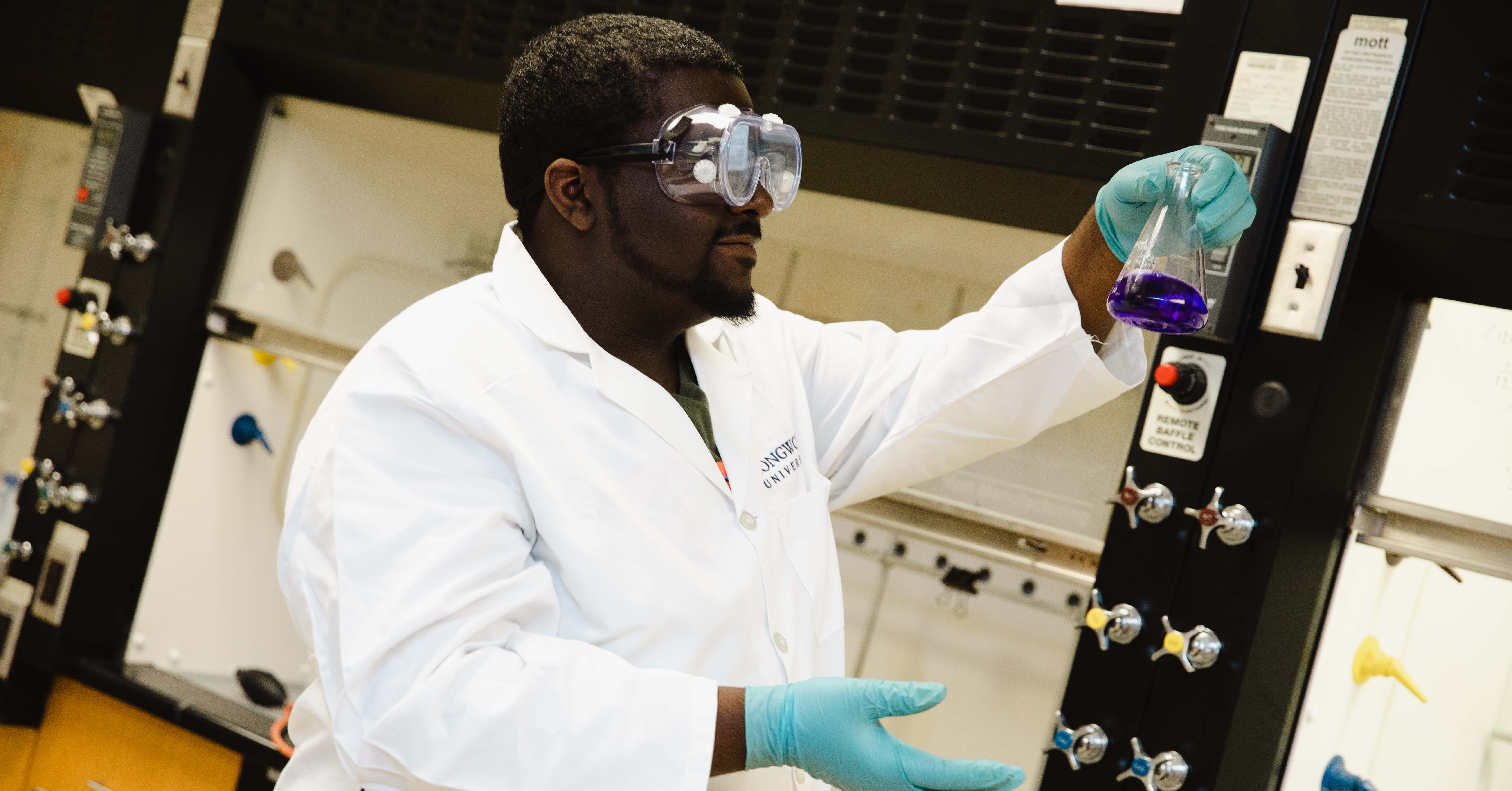 Antonio Harvey ‘26 - A student wearing safety goggles and gloves works in a chemistry lab. He holds up a flask containing a purple liquid. He stands beside a lab bench with gas valves and equipment. The scene shows hands-on scientific experimentation.