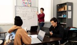 Students working in the QR center - A student stands at a whiteboard explaining math equations. Two classmates sit at a table working on laptops. Formulas and calculations fill the board behind the presenter. The scene shows collaborative problem-solving in a study session.
