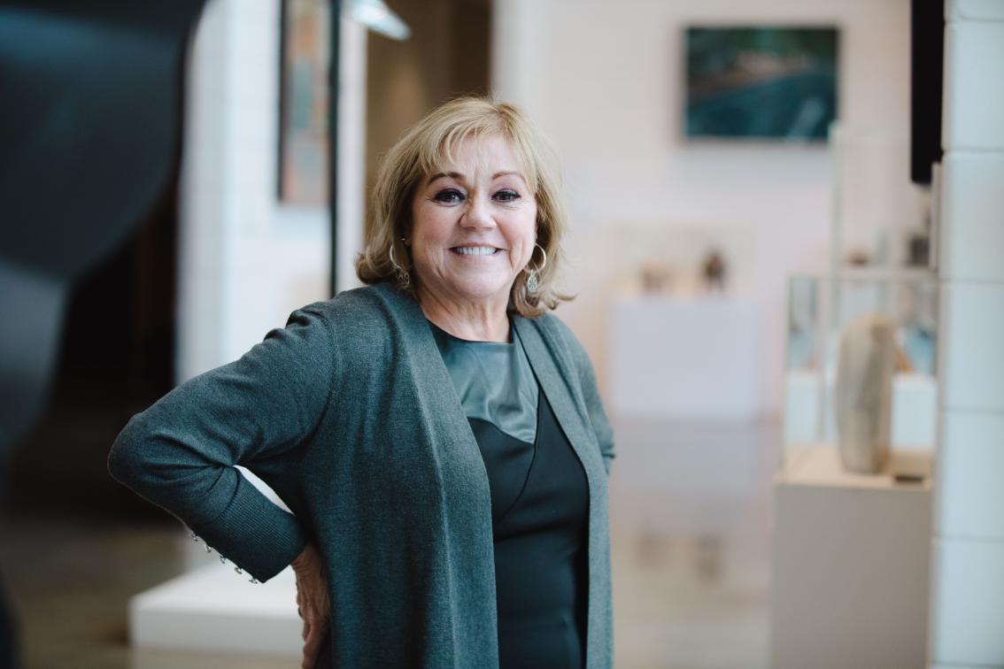 Ronda Landis Scarrow ’80 - A woman smiles while standing in a bright gallery space. She wears a dark green dress with a matching cardigan. Her hand rests on her hip as she faces the camera. Artwork and sculptures are softly visible in the background.