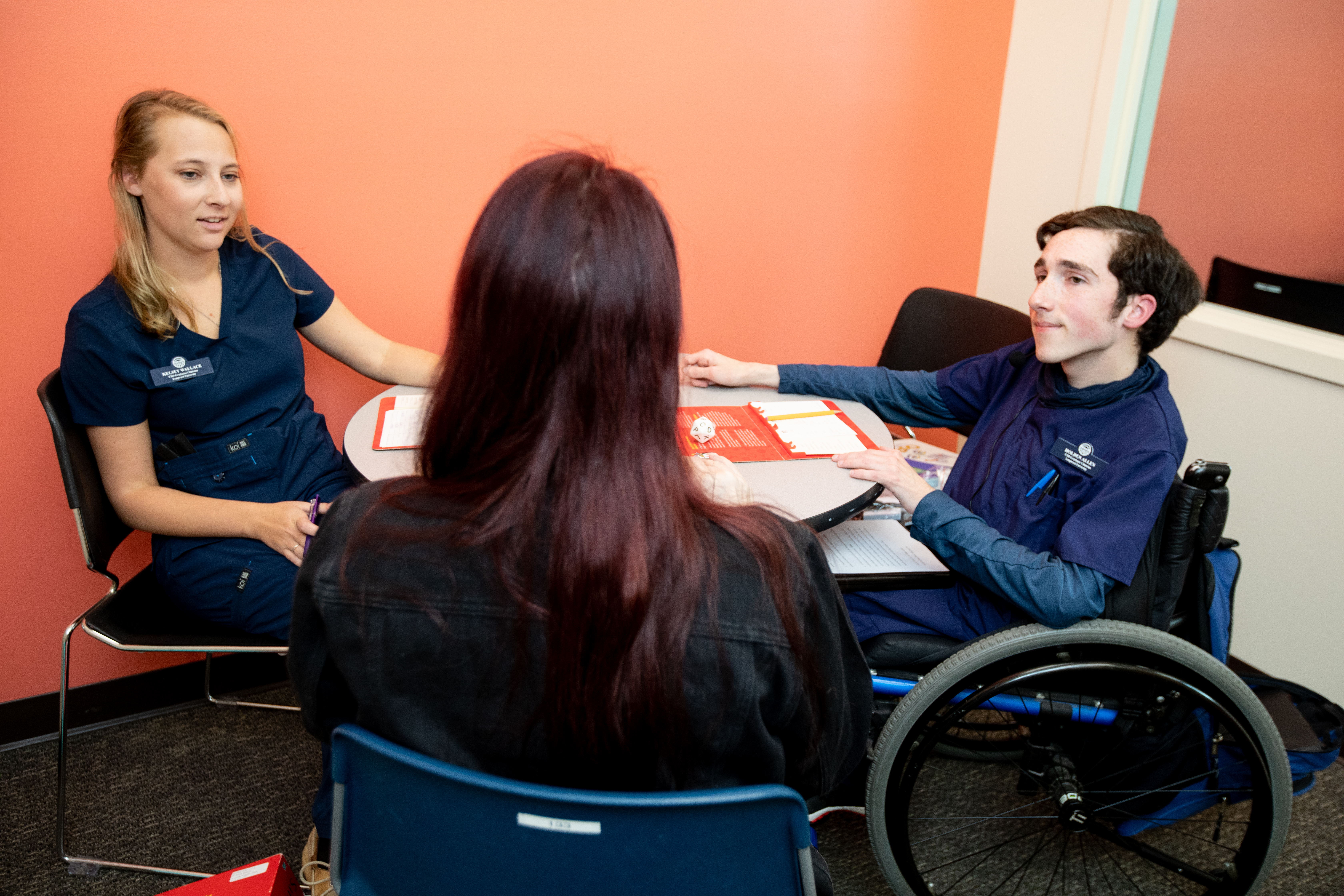 Communication sciences and disorders graduate students work with a client - Two healthcare students in navy scrubs sit at a small table speaking with a visitor. One student uses a wheelchair while reviewing paperwork and a red folder. The visitor sits with her back to the camera in a campus office setting. They appear engaged in a focused, professional conversation.