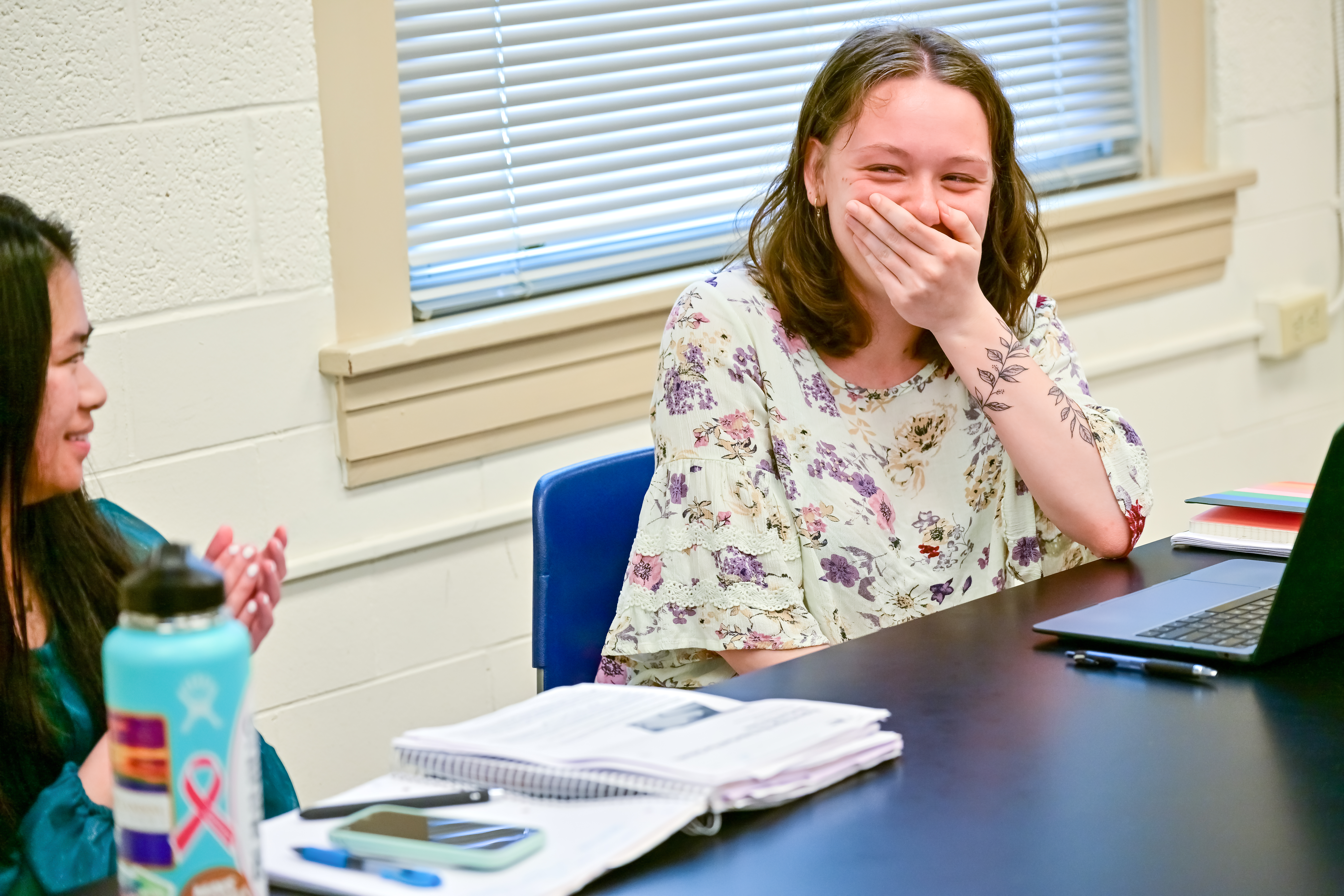 Emily Robertson '24 reacts to the news that she is Longwood's first-ever Truman Scholar. - A student laughs while sitting at a table with a laptop and notebooks. She covers her mouth, reacting to something amusing. Another student sits nearby with a water bottle and papers. The setting appears to be a casual classroom discussion.