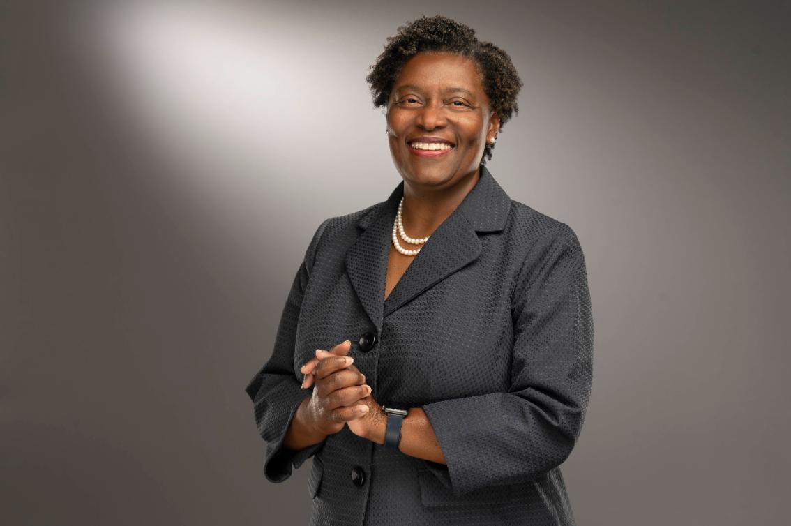 Dr. Tamara Brown ’89 - Professional portrait of a smiling woman with short natural hair. She wears a dark textured blazer and a pearl necklace. Her hands are clasped in front of her. The background is a soft gray studio backdrop.