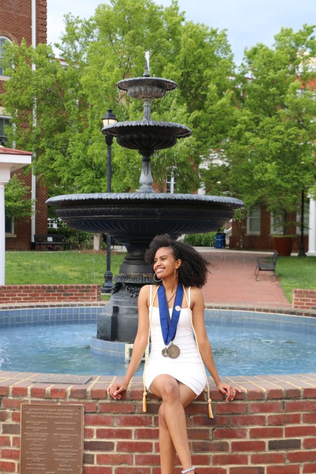 Tinsae Alem ’23 - Graduate wearing medals and honor cords sits on a brick fountain ledge. She smiles while posing in front of a tiered campus fountain. Red brick buildings and green trees fill the background. Outdoor portrait taken on a sunny day.