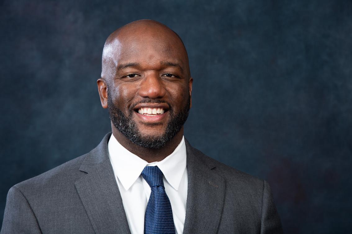 Troy Austin - Professional headshot of a smiling man with a trimmed beard. He wears a gray suit jacket, white dress shirt, and blue tie. The portrait is set against a dark blue studio background. He faces the camera with a confident expression.