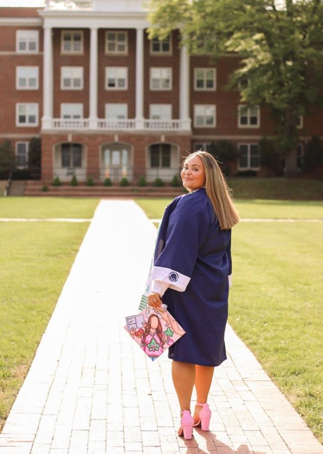 Aryanna Colvin ’23 - A graduate in a navy cap and gown walks along a campus pathway. She looks back and smiles while holding a decorated graduation cap. A red brick academic building with white columns stands in the background. The photo captures a celebratory moment on a sunny day.