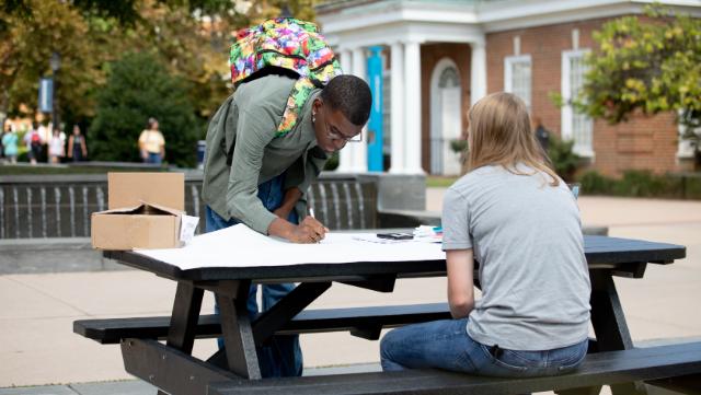 Several initiatives like last fall's Voter Registration Drive, led by a joint effort between the College Democrats and College Republicans, have led Longwood to be named a Voter Friendly Campus. - A student leans over a picnic table to sign a large sheet of paper outdoors. Another student sits across from him with markers and supplies spread out on the table. A colorful backpack rests on his shoul