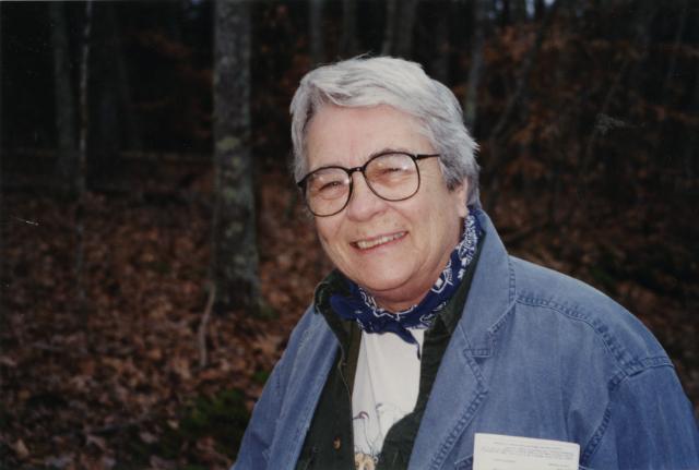 Dr. Carolyn Wells - Older woman with short gray hair and glasses smiling outdoors. She wears a denim jacket and patterned neck scarf. Autumn leaves and trees fill the wooded background. Candid portrait taken in natural light.
