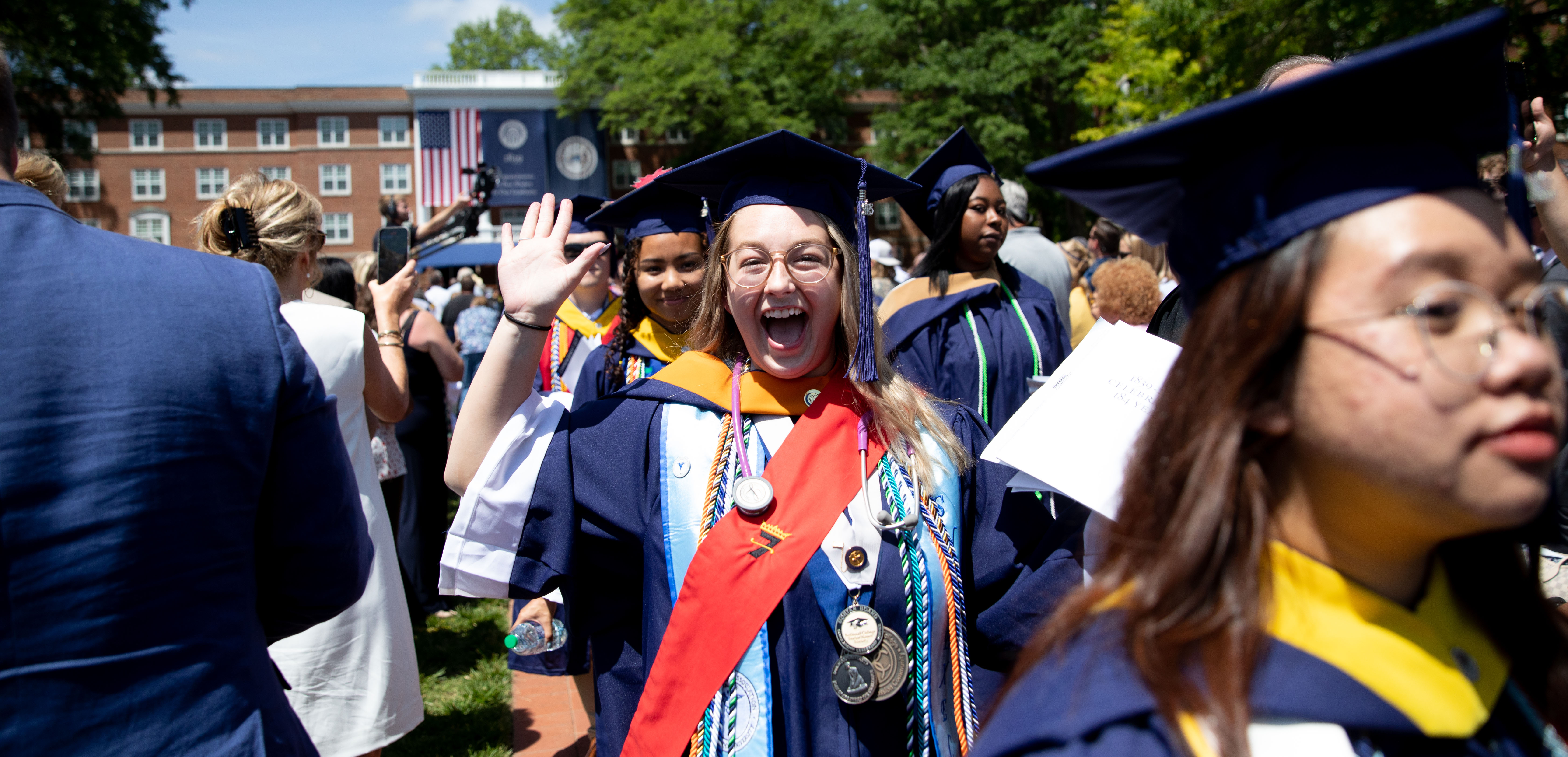 Excited student during the Commencement ceremony - A graduate in cap and gown smiles widely and waves during commencement. She wears honor cords and medals over her academic regalia. Other graduates and guests fill the lawn in front of a campus building with an American flag. The image captures a joyful moment during graduation festivities.