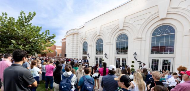 Crowd awaits the Joan Perry Brock Center ribbon cutting. The building is in the background. - A large crowd gathers outside a white campus building with tall arched windows. People stand facing a podium set up near the entrance for a formal event. The building façade features decorative architectural details and lantern-style lights. Trees and blue sky frame the outdoor ceremony scene.