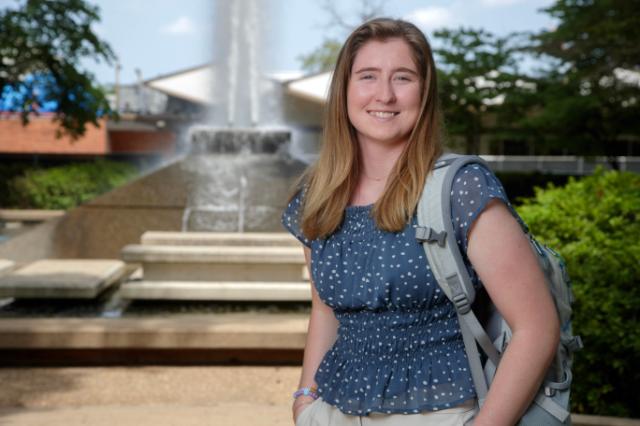 Gracie Hess '24, credit: University of Arkansas at Little Rock - A student stands smiling in front of a campus fountain on a sunny day. She wears a blue polka dot blouse and a gray backpack over one shoulder. Water cascades behind her with trees and campus buildings in the background. The scene captures a bright, welcoming campus setting.