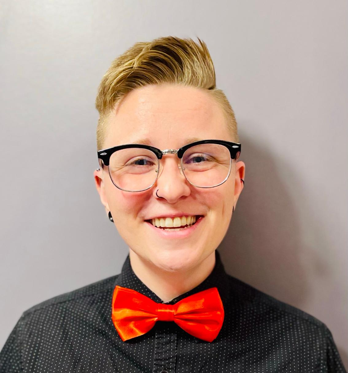Tiarrah “T” Crouch Parker ’17 - Headshot of a smiling person with short blond hair and black-framed glasses. They wear a black button-down shirt with a subtle pattern. A bright red bow tie stands out against the dark shirt. The portrait is set against a neutral gray background.
