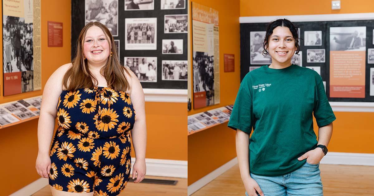 Hailey Belote ’23 and Gabrielle Bustillos ’24 - Two students stand smiling in a gallery-style campus exhibit space. One wears a sunflower-patterned dress, the other a green T-shirt and jeans. Historic photographs and display panels line the orange walls behind them. The portrait highlights student presence in a campus history setting.