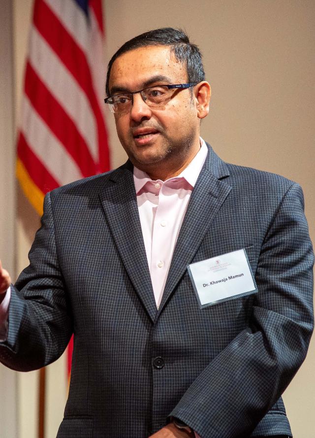Dr. Khawaja Mamun - A man in a dark blazer and glasses speaks during a formal event. He wears a name badge that reads “Dr. Khawaja Mamun.” An American flag stands in the background behind him. He gestures with one hand while addressing the audience.