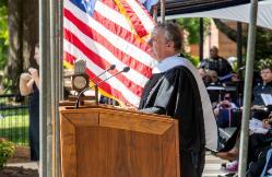 Author and columnist John Feinstein was the keynote speaker at Longwood's 2023 undergraduate Commencement ceremony - University official in academic regalia speaks at a wooden podium outdoors. An American flag hangs prominently behind him. Audience members sit under a tent during the ceremony. Trees and campus buildings are visible in the background.