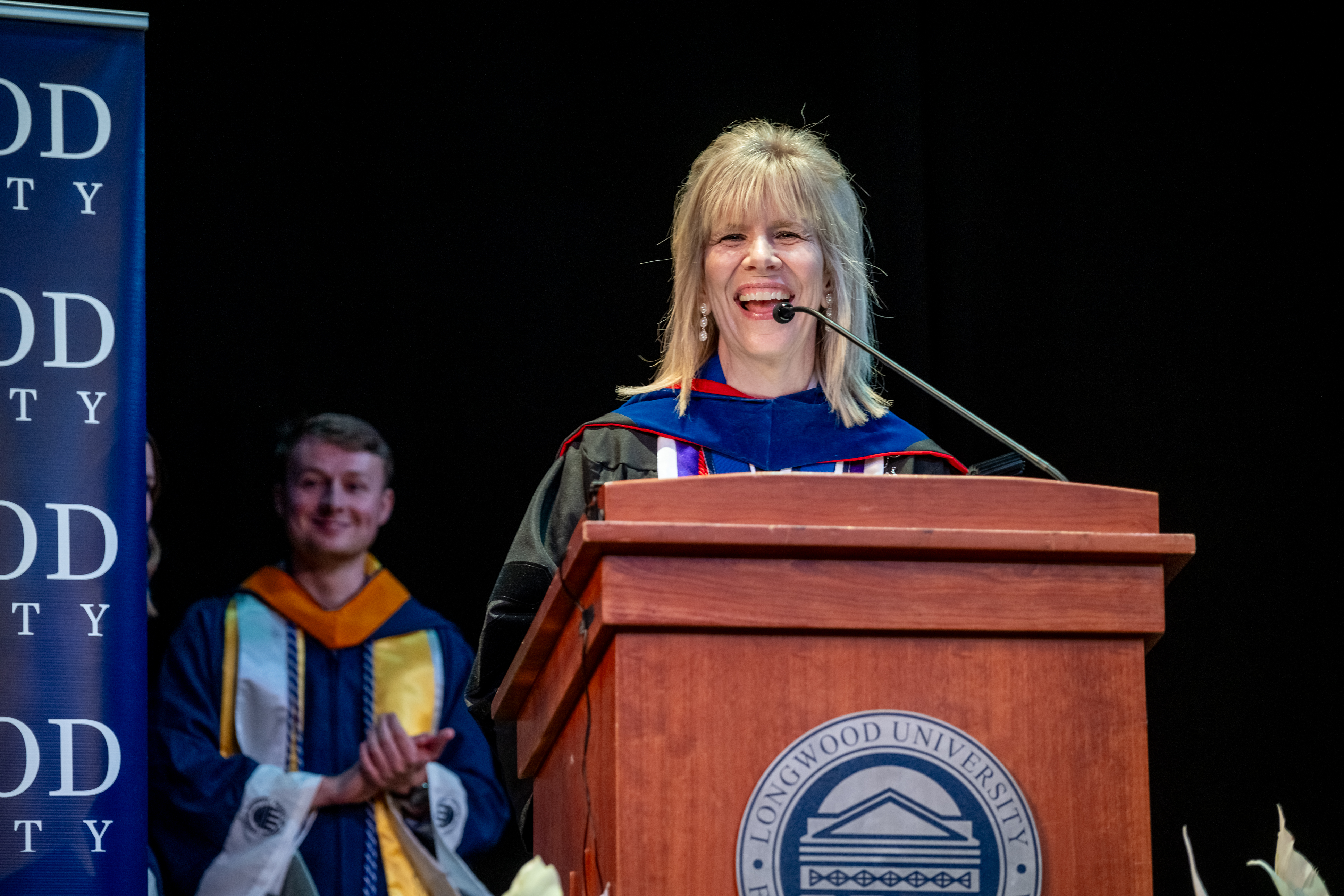 Dr. Kim Little - Woman in academic regalia smiles while speaking at a podium with the Longwood University seal. She stands on a stage in front of a microphone during a formal ceremony. A student in graduation attire applauds in the background. A Longwood University banner is visible beside the stage.