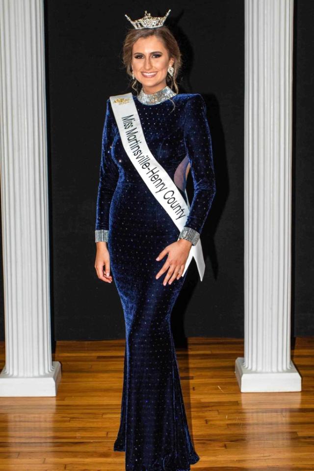 Jenna Adams ’23 - Woman wearing a crown and sash stands between white columns. Her sash reads “Miss Martinsville-Henry County.” She wears a long navy velvet gown with sparkling details. Formal portrait taken on a wooden stage floor.