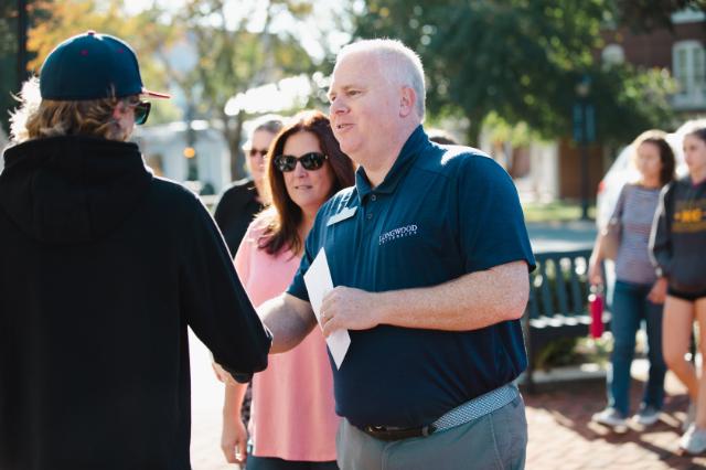 Jason “Ferg” Ferguson talks to family members at an admissions event - A Longwood University staff member greets a visitor outdoors. He wears a navy polo with the university logo and a name badge. A woman stands nearby as other guests walk in the background. The scene takes place on a sunny campus walkway.