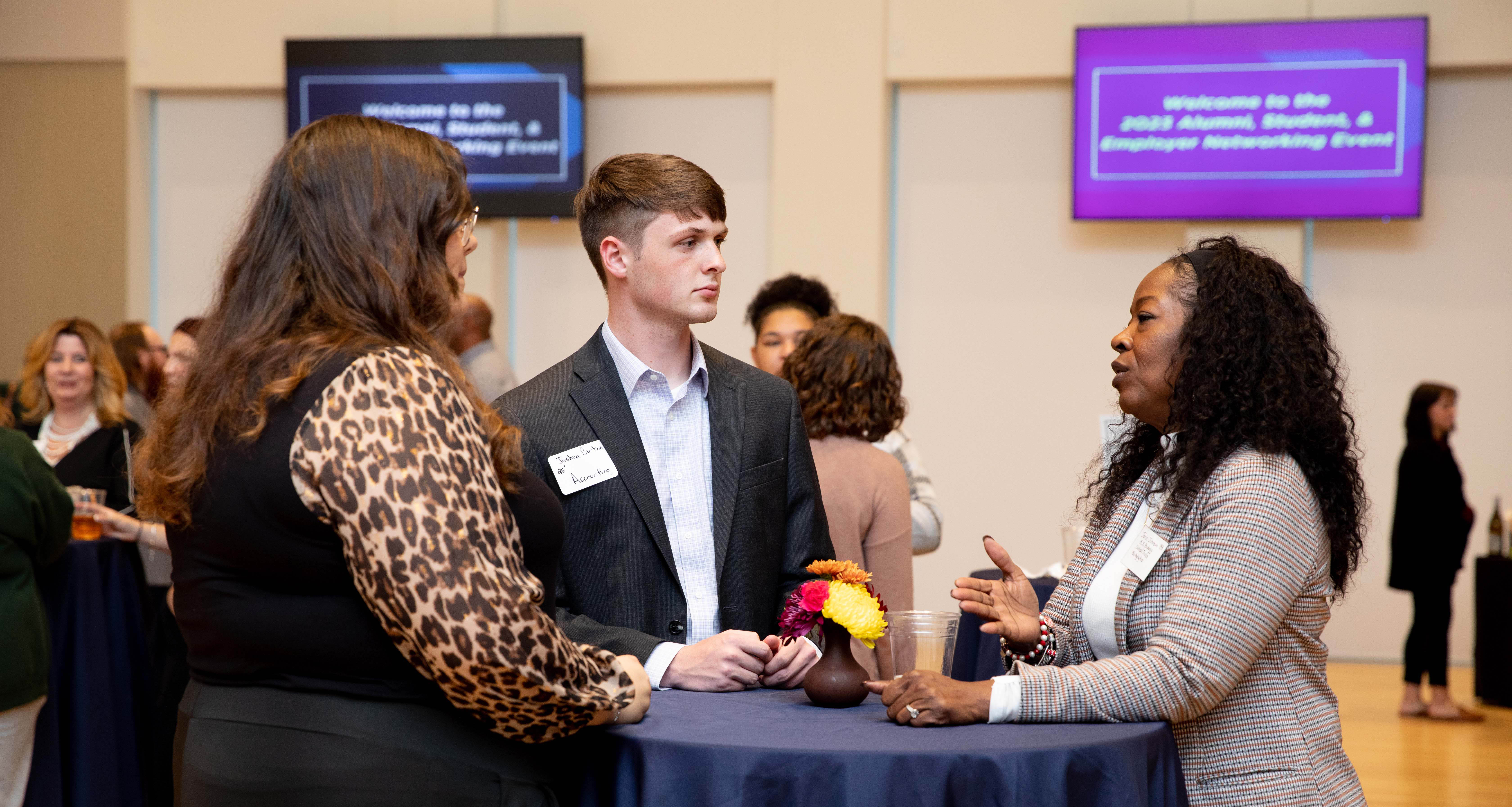 Students engage with professionals and alumni in a variety of a fields at networking events hosted by the Center for Career Success. - Three attendees talk around a table at an alumni networking event. A student in a suit listens as two professionals engage in conversation. A small vase of colorful flowers sits on the table between them. Event welcome screens glow in the background above the crowd