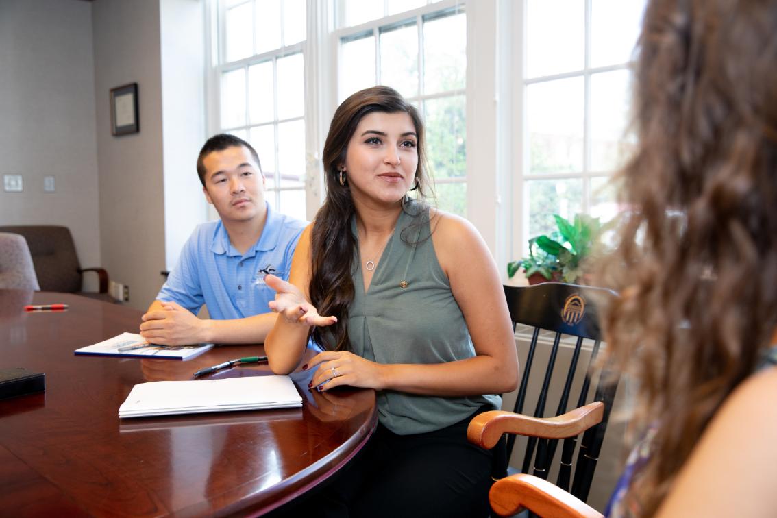 - Three students sit around a round wooden table in a bright room with tall windows. A woman in a green sleeveless blouse gestures as she speaks, papers and a pen in front of her. A man in a blue polo shirt listens with a notebook open on the table. Another student is partially visible in the foreground, facing the group.