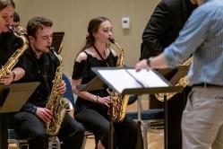 Music students playing with sheet music in the foreground - Student musicians play saxophones during a concert performance. They sit in a row reading from music stands while a conductor leads in the foreground. The performers wear black concert attire. The scene takes place indoors in a recital or performance hall.