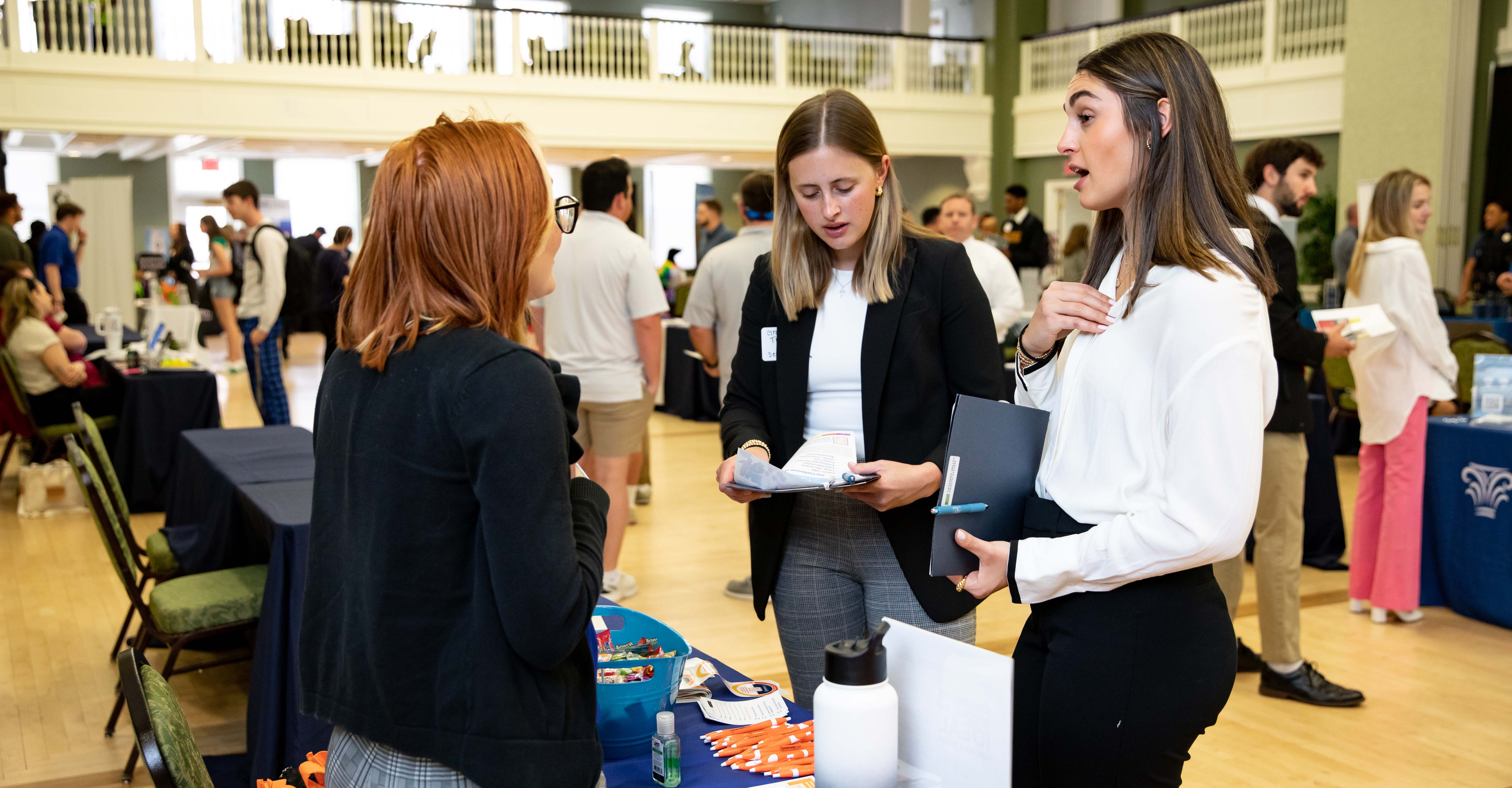 Students speaking to an employer at a career fair - Three students talk at a table during a campus career fair. Two hold folders and review printed materials while speaking with a representative. Recruiter tables and attendees fill the bright, open hall in the background. Promotional items and brochures are spread across the table in the foreground.