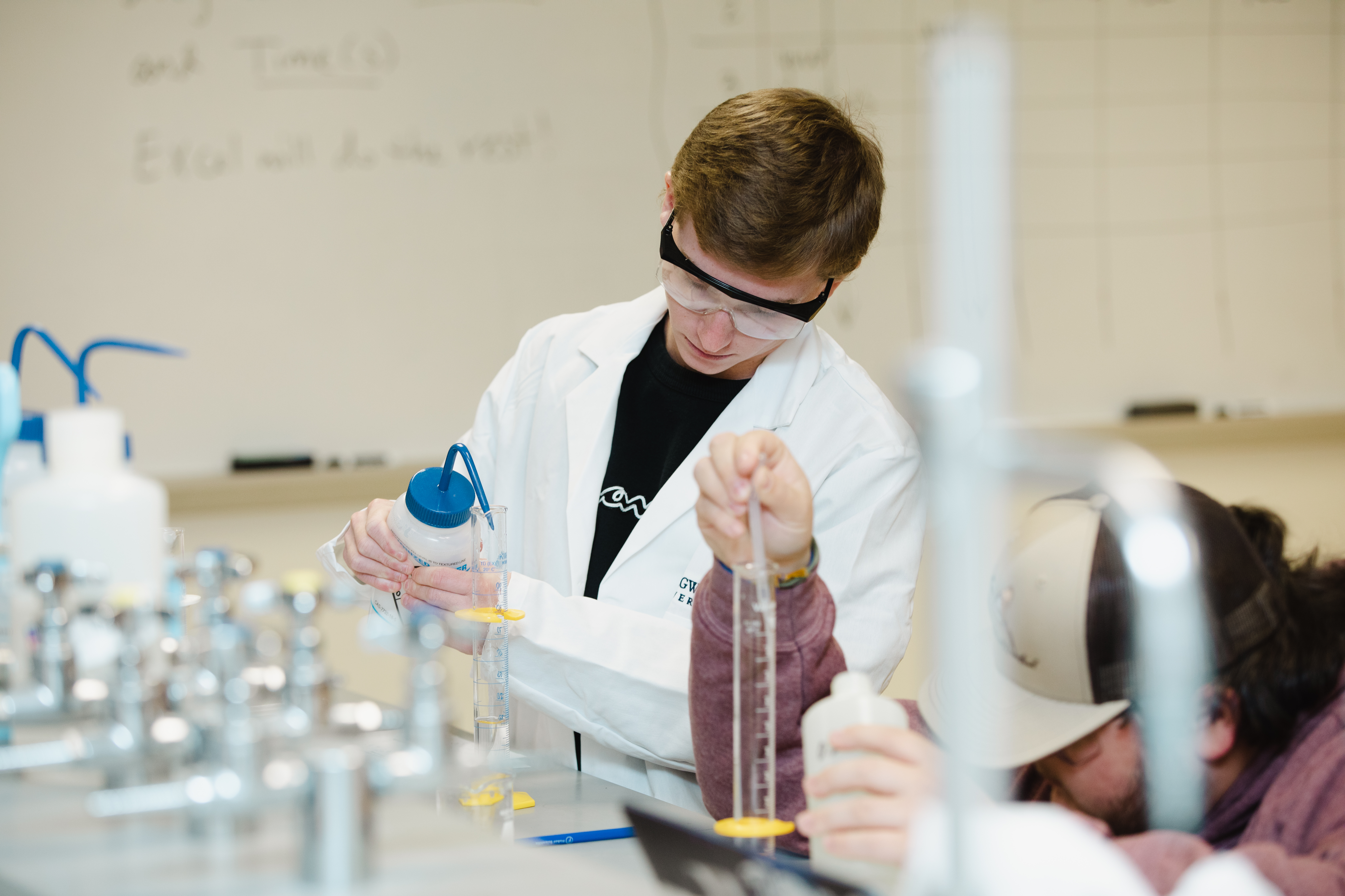 Students working in a lab at Longwood - Student in a lab coat and safety goggles measures liquid in a graduated cylinder. Another student uses a pipette beside him. They work at a laboratory bench with glassware and equipment. A whiteboard is visible in the background.