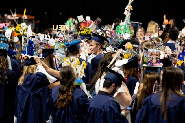 Student in the midst of the sea of caps at Convocation 2024 - Graduates in navy caps and gowns gather indoors at commencement. Many wear decorated mortarboards featuring photos, flowers, and colorful signs. Students stand close together, talking and taking photos. The celebratory scene captures the excitement of graduation day.