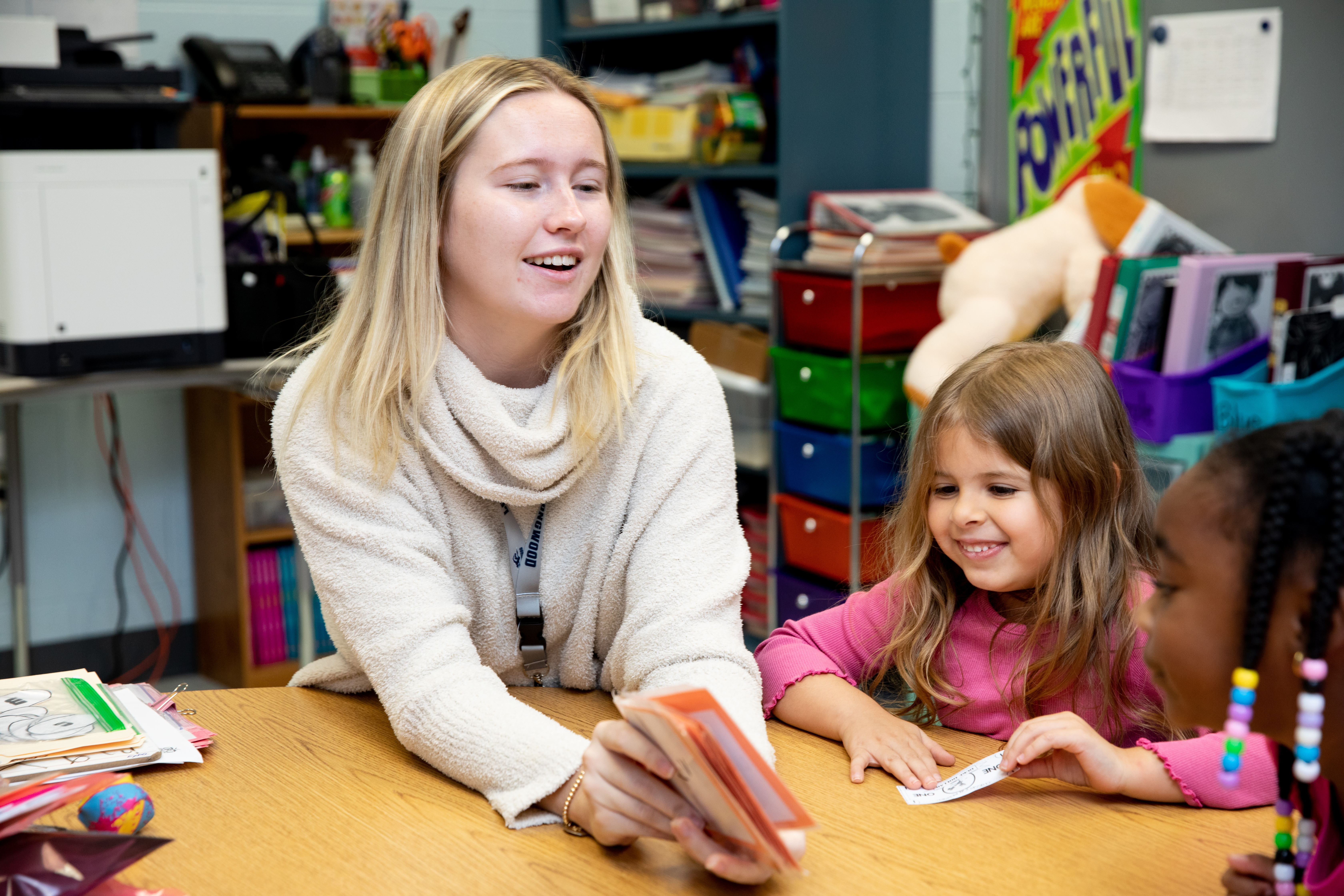 Student teacher in classroom with some elementary school students - Teacher sits at a classroom table holding flashcards while working with two young children. One child smiles as she looks at the card, while another child listens from the side. Colorful bins, books, and classroom supplies fill the shelves in the background. The small group appears engaged in an early learning activity.