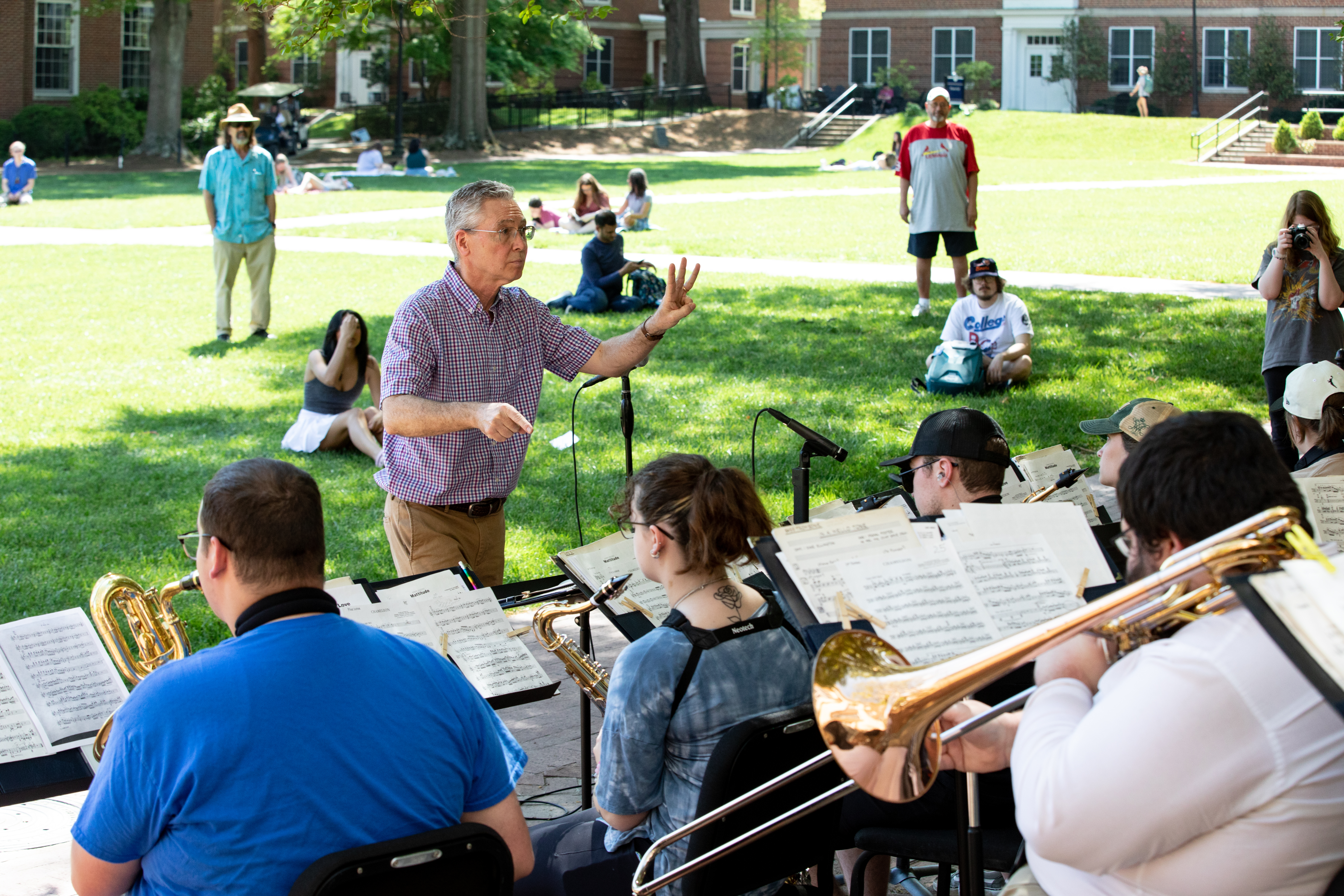 Dr. Charlie Kinzer conducts the Jazz Ensemble performing on campus - Music professor conducts a student ensemble performing outdoors on campus. Brass players read sheet music while seated under leafy trees. Audience members sit and stand on the lawn near brick academic buildings. Sunny scene captures a live performance in a campus courtyard.