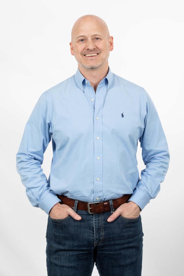 Dr. Eric Hodges, associate professor of political science - Man stands against a light gray studio background with hands in his pockets. He wears a light blue button-down shirt and dark jeans. He smiles directly at the camera. Professional portrait with a relaxed, approachable demeanor.